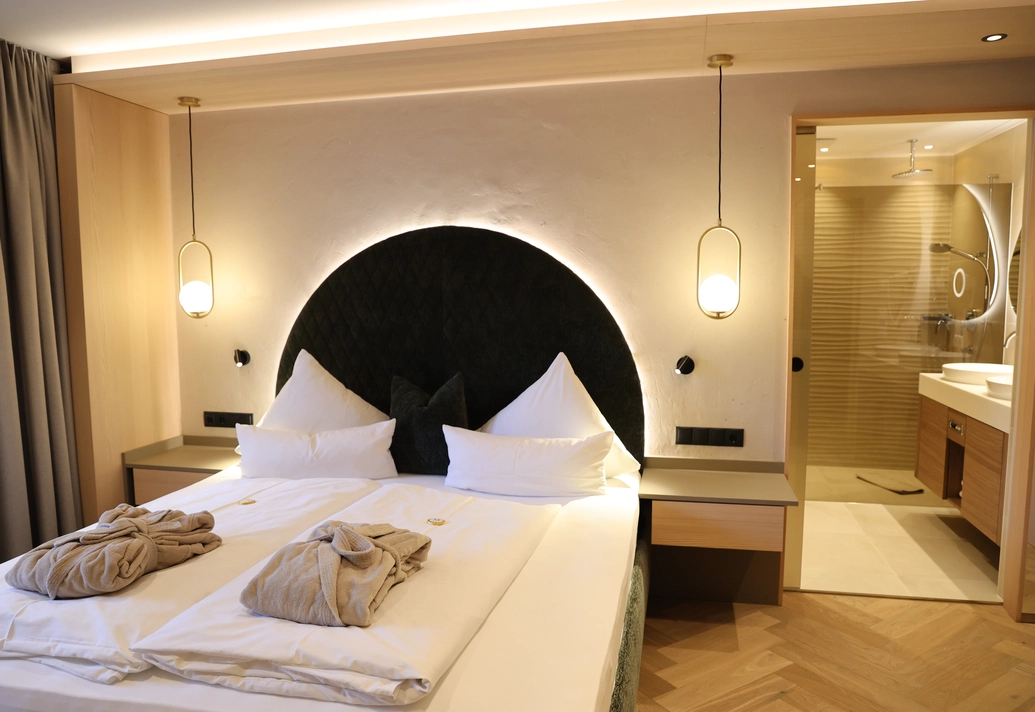 Großes Doppelbett mit rundem, beleuchtetem Kopfteil und Blick in das Badezimmer im Metzgerwirt Zillertal.